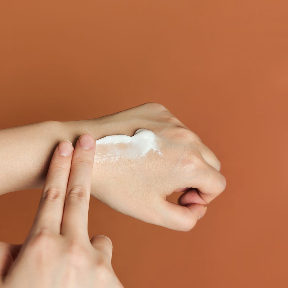 Main avec crème sur le bras. La crème est étalée avec deux doigts. Fond brun.