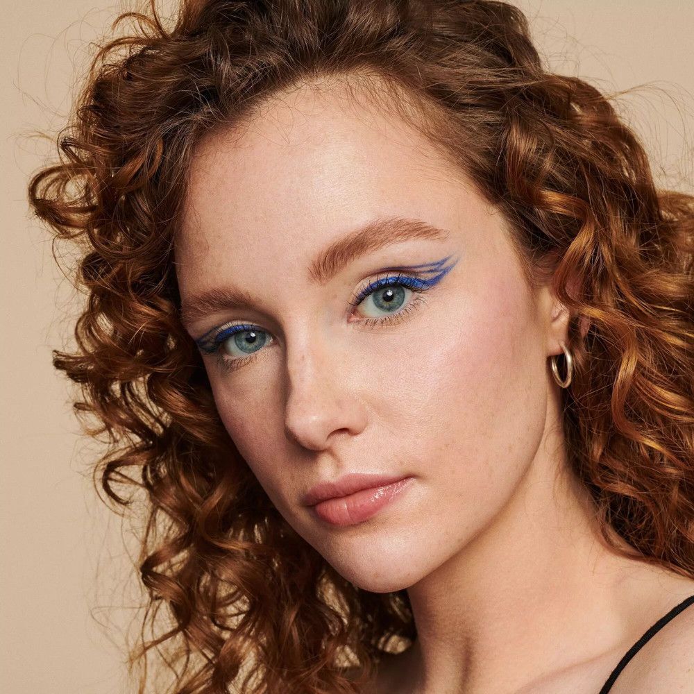 Close-up van een gezicht met een blauwe eyelinerlijn en wenkbrauwen. De vrouw heeft krullend haar.