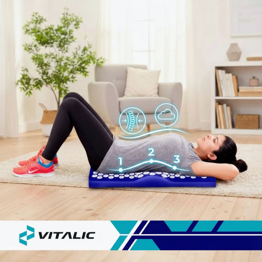 Persoon ligt op blauwe acupressuurmat. VITALIC logo. Omgeving: tapijt, bank, plant.