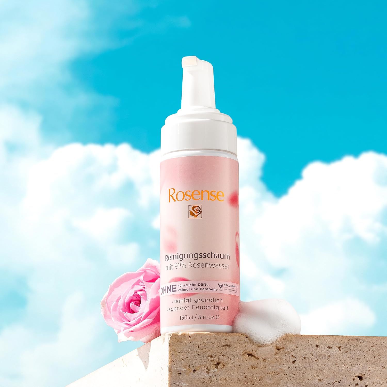 Flacon de mousse nettoyante Rosense, étiquette rose, devant un ciel bleu. Rose et mousse à côté. Texte: 91% eau de rose, végétalien.