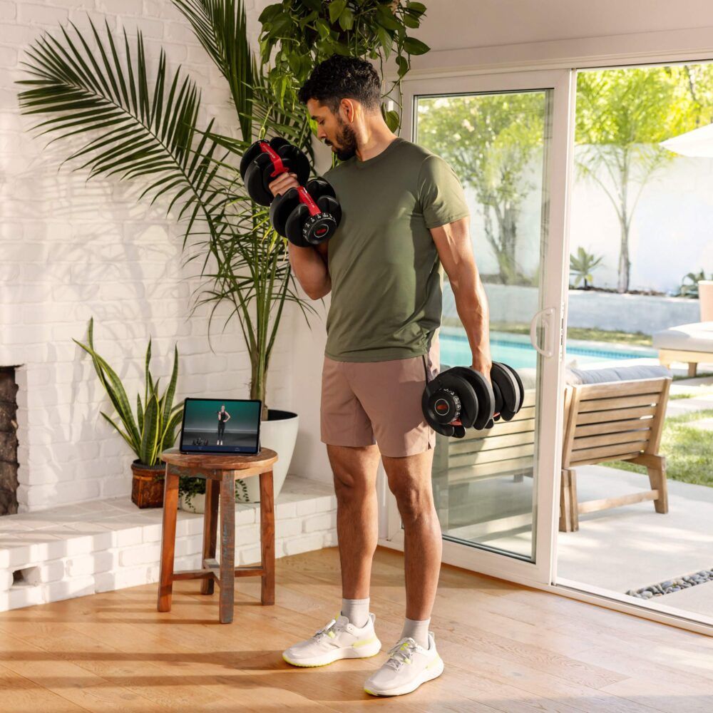 Man traint met twee dumbbells. Tablet met trainingsvideo. Interieur met meubels en planten.