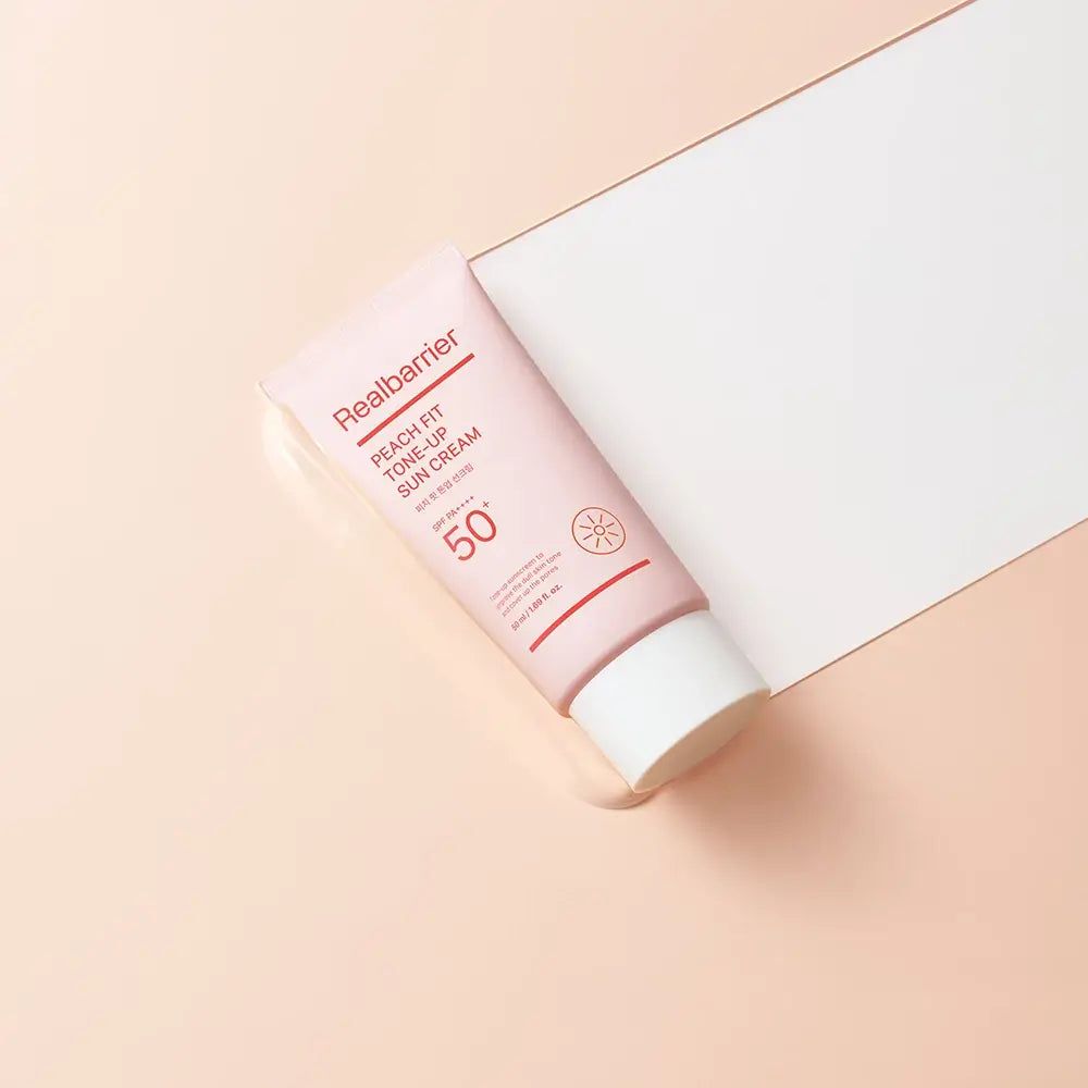 Tube rose avec bouchon blanc. Inscription: Realbarrier Peach Fit Tone-Up Sun Cream LSF 50+ PA++++. Symbole de soleil.
