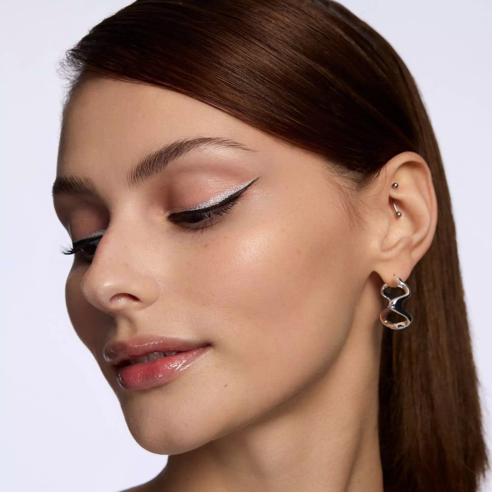 Portrait d'une femme avec un trait d'eye-liner argenté. Yeux ouverts, piercing, boucles d'oreilles. Peau claire, lèvres roses.