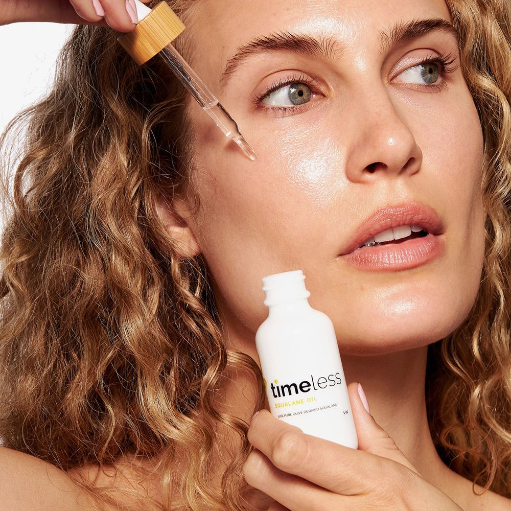 Vrouw met krullen die olie met een pipet op haar gezicht aanbrengt. Witte fles met productnaam en ingrediënten.