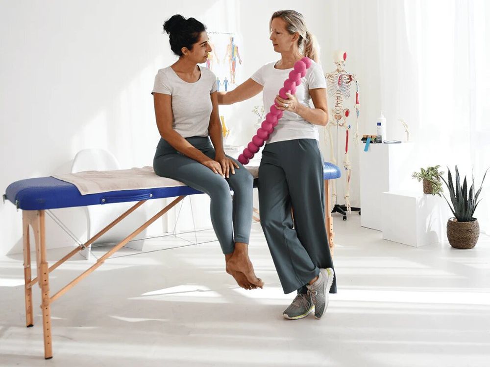 Vrouw houdt roze SPINEFITTER® vast. Twee vrouwen in behandelkamer. Eén zit op een bed.