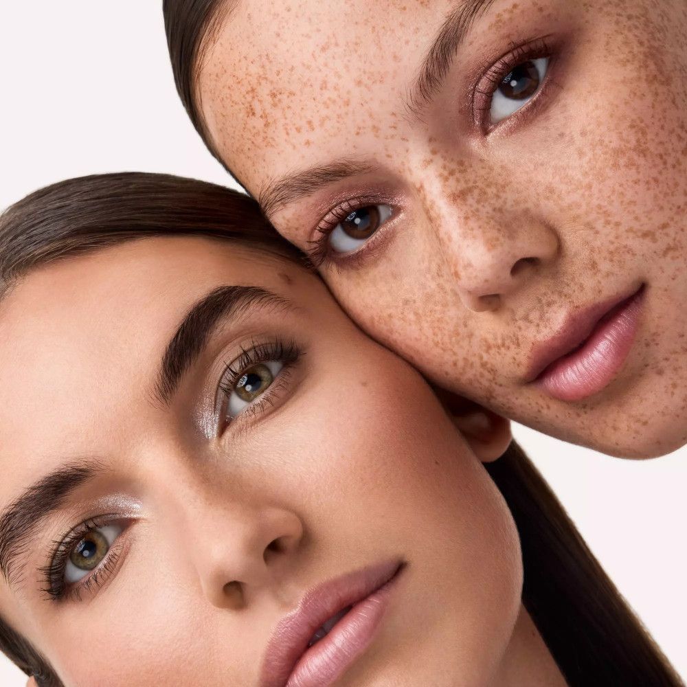Gros plan de deux femmes. Une avec des taches de rousseur, l'autre avec un teint clair. Les deux portent du maquillage des yeux. Fond clair.