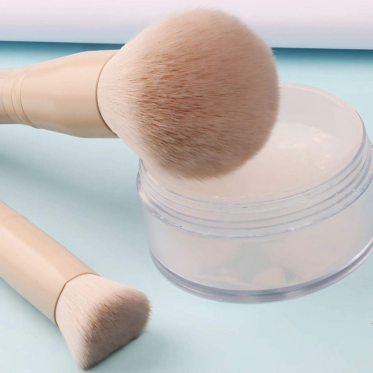 Deux pinceaux de maquillage à manches beiges clairs à côté d'un récipient transparent contenant de la poudre blanche.