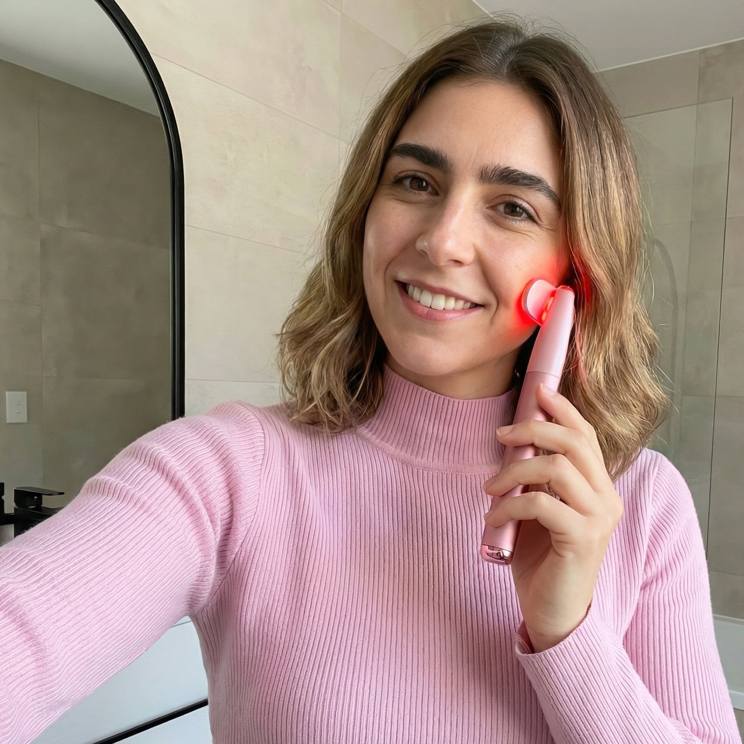 Vrouw houdt een roze gezichtsmassager vast. Het apparaat heeft een langwerpig lichaam en een massagekop.