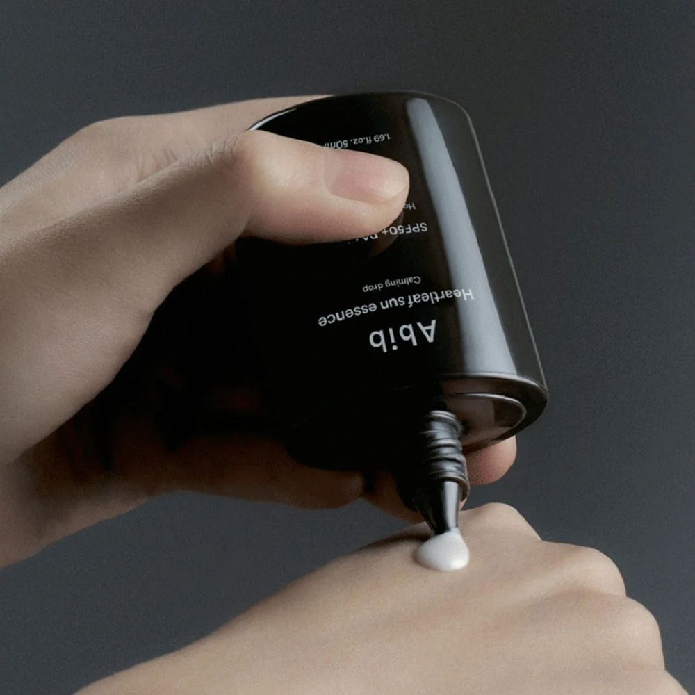 Hand die crème uit een zwarte fles drukt. Opschrift: Abib Heartleaf Sun Essence Calming Drop SPF50+ PA++++.