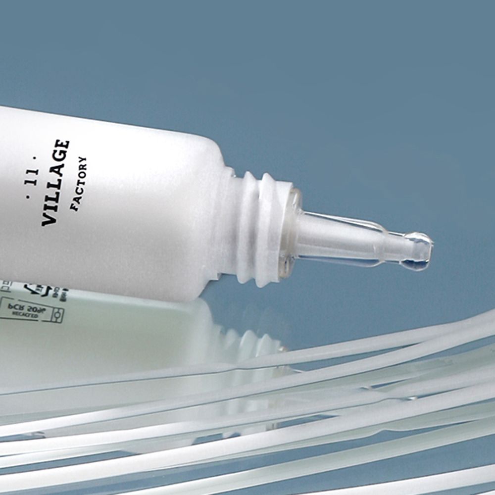 Witte tube, bruine sluiting, transparante punt. Zwarte tekst: VILLAGE 11 FACTORY. Op glad oppervlak.
