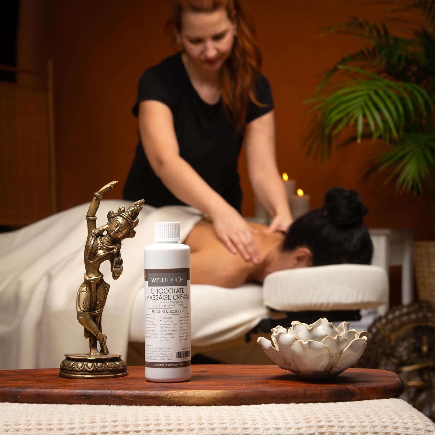 Witte fles Welltouch Chocolate Massage Cream. Opschrift: Chocolate Massage Cream. Bruine dop, wit etiket. Vrouw krijgt massage.