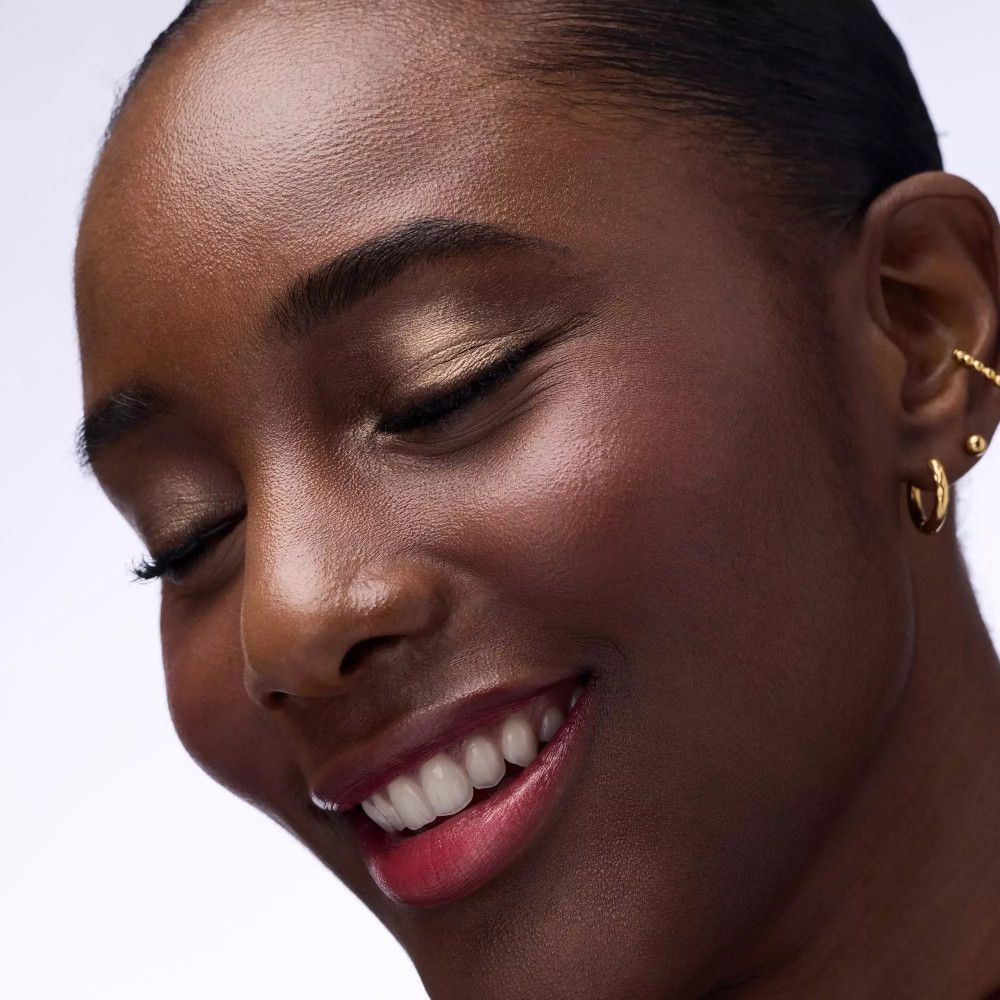 Gros plan d'une femme avec fard à paupières doré. Elle sourit. Bijoux d'oreille dorés visibles. Rouge à lèvres et fard à joues.