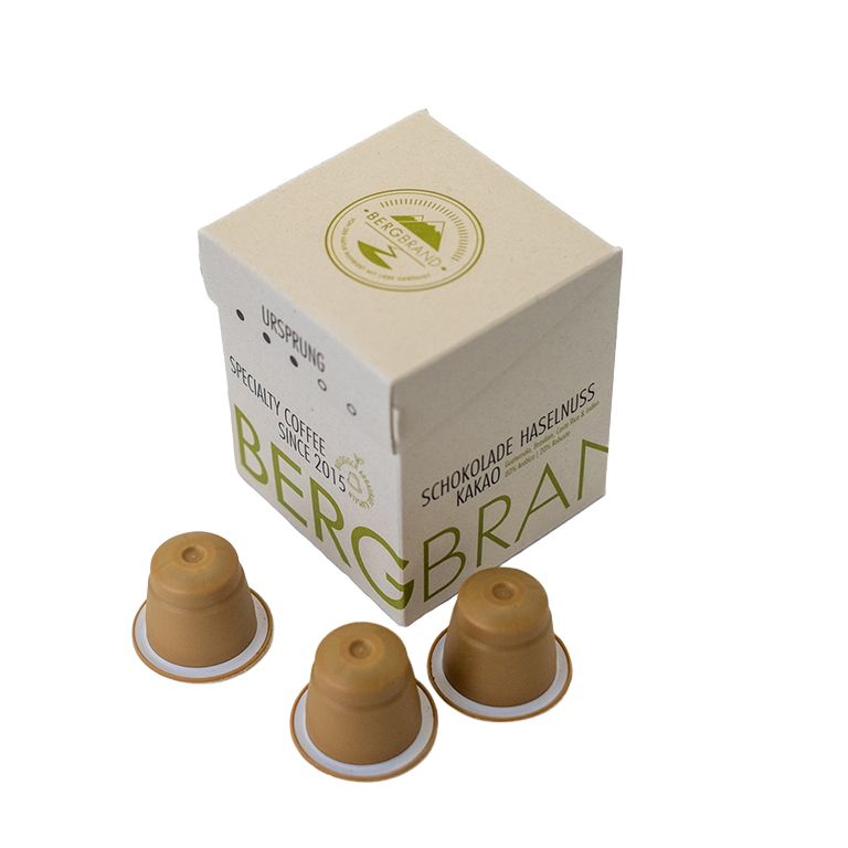 Koffiecapsules en verpakking. Opschrift: BERGBRAND, Ursprung, Schokolade Haselnuss Kakao. Capsules bruin, verpakking beige.