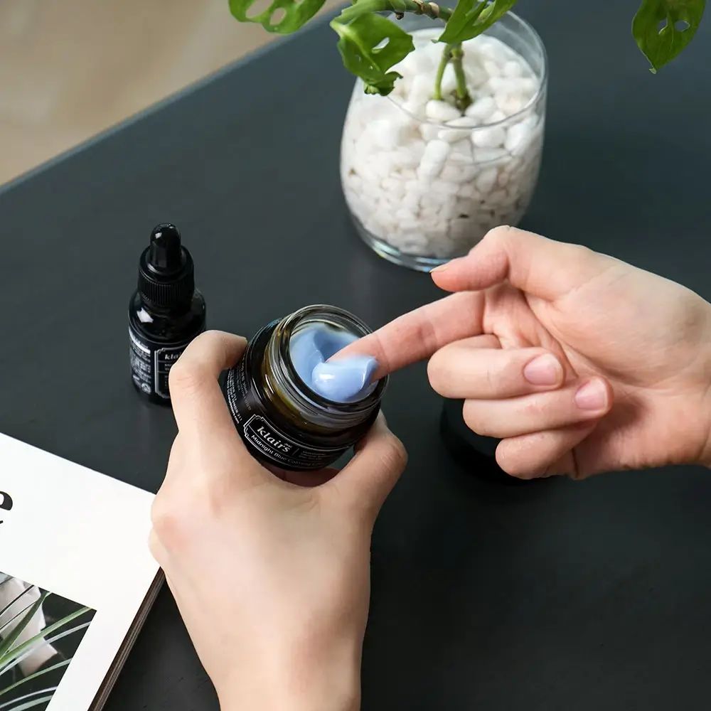 Handen houden een open potje met blauwe crème vast. Daarnaast een flesje met zwart etiket.