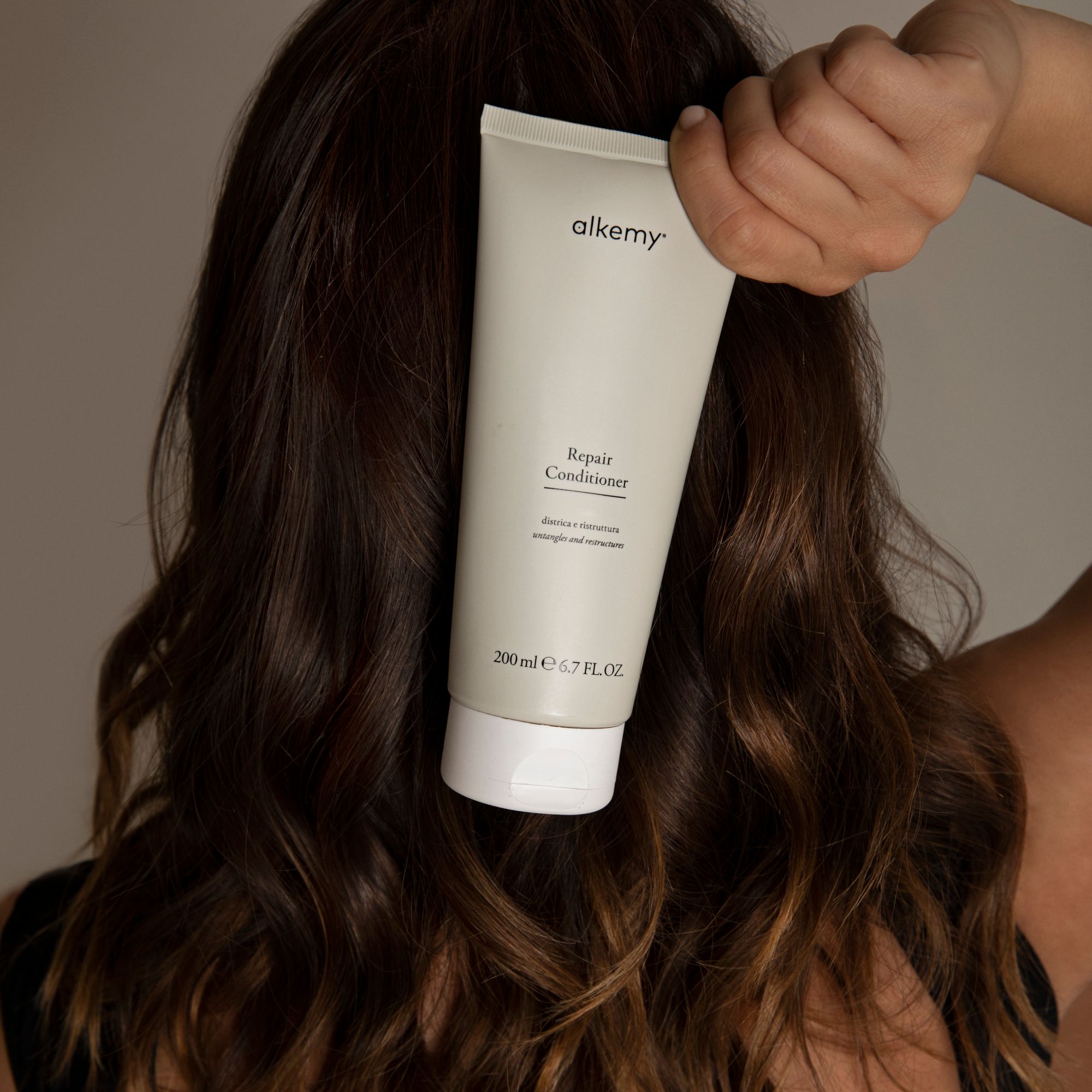 Un tube de revitalisant réparateur Alkemy, 200 ml, tenu derrière les cheveux d'une personne.
