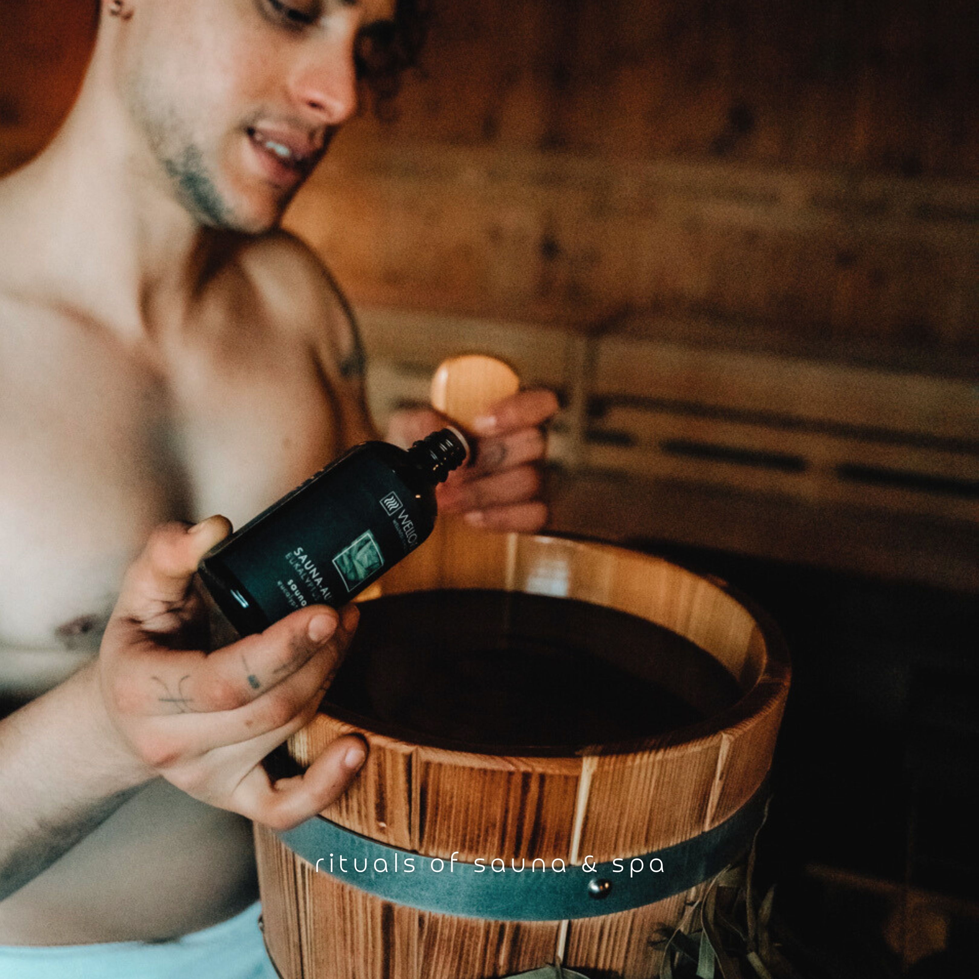 Homme dans un sauna, tenant une bouteille et un seau en bois. Inscription : Sauna-Aufguss.