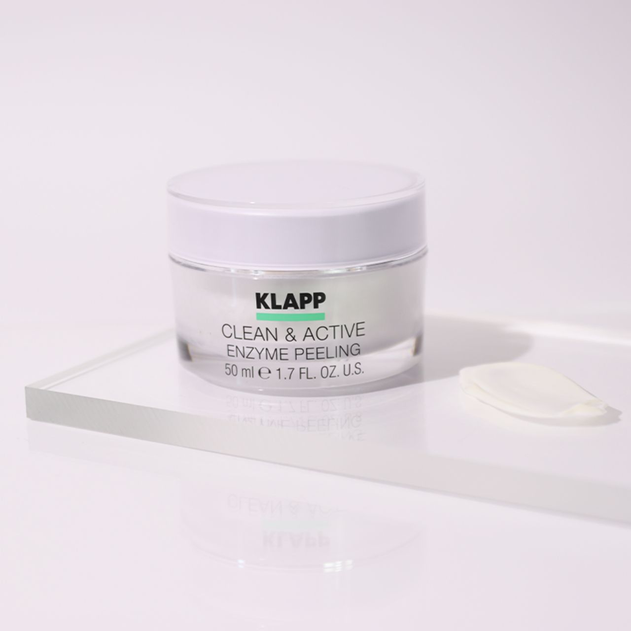 Pot transparent Klapp Clean & Active Enzyme Peeling avec couvercle blanc. Sur une plaque transparente. À côté, une goutte.