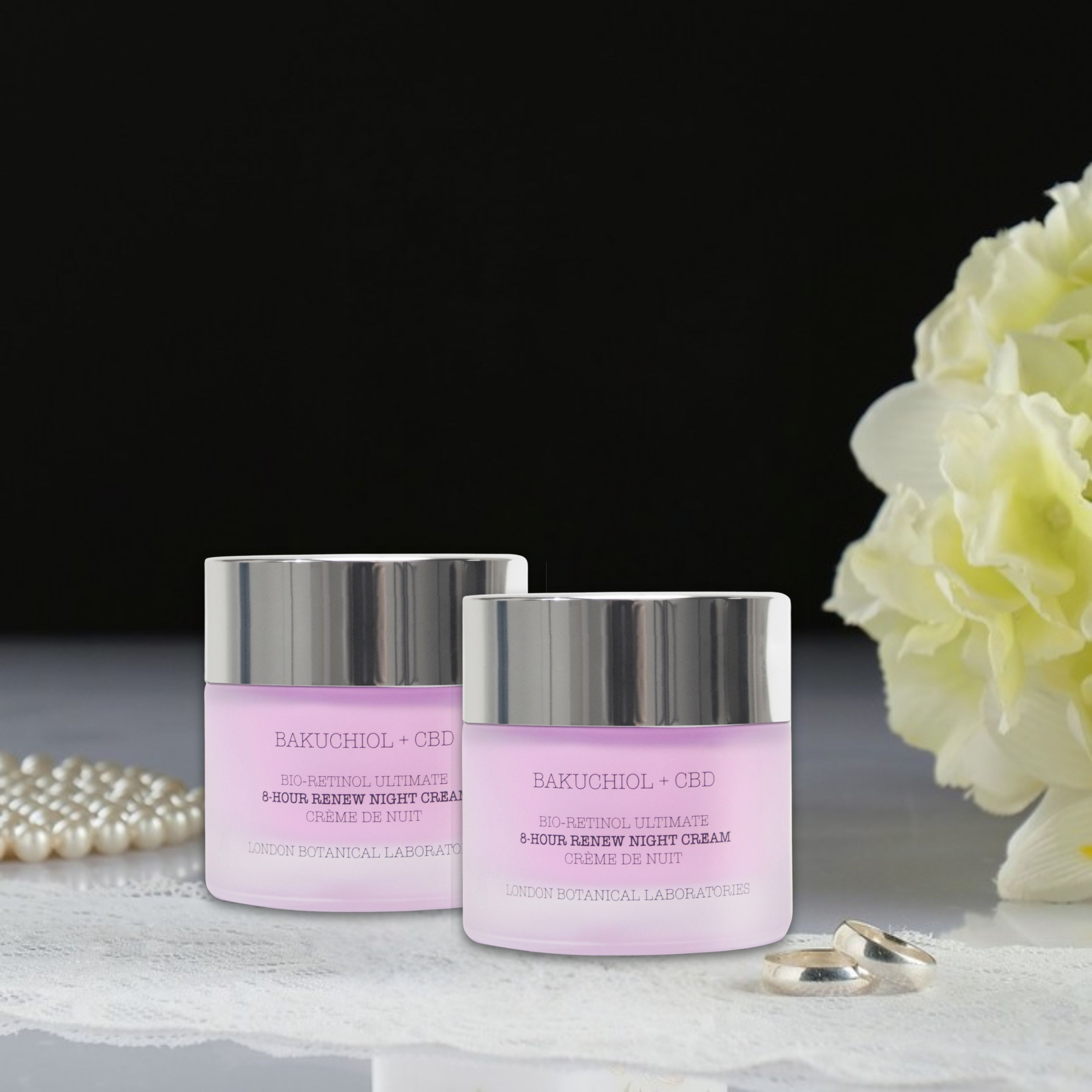 Twee potten nachtcrème met zilveren deksels. Opschrift: Bakuchiol + CBD. Bloemen en sieraden op de achtergrond.