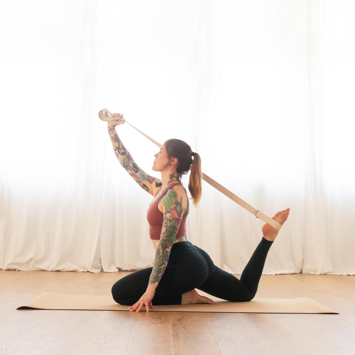 Femme tatouée utilisant une sangle de yoga dans une pose de yoga. Sangle beige, anneaux métalliques.
