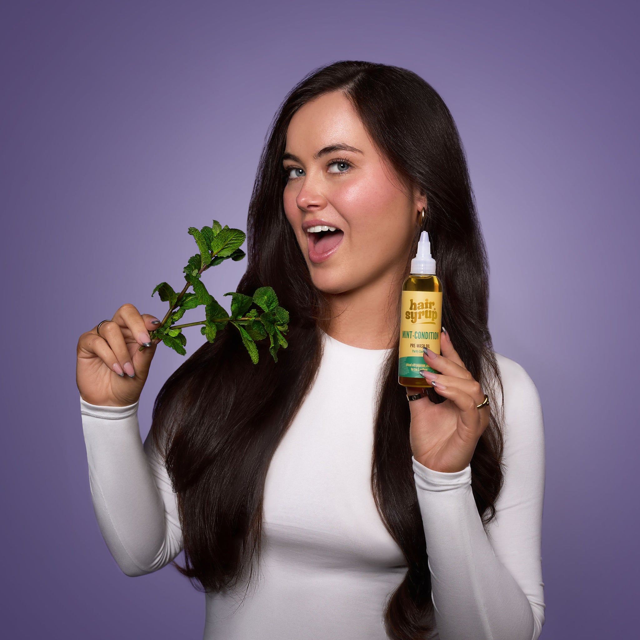 Vrouw met lang haar houdt fles "Hair Syrup Mint Condition" en takje munt vast. Paarse achtergrond.