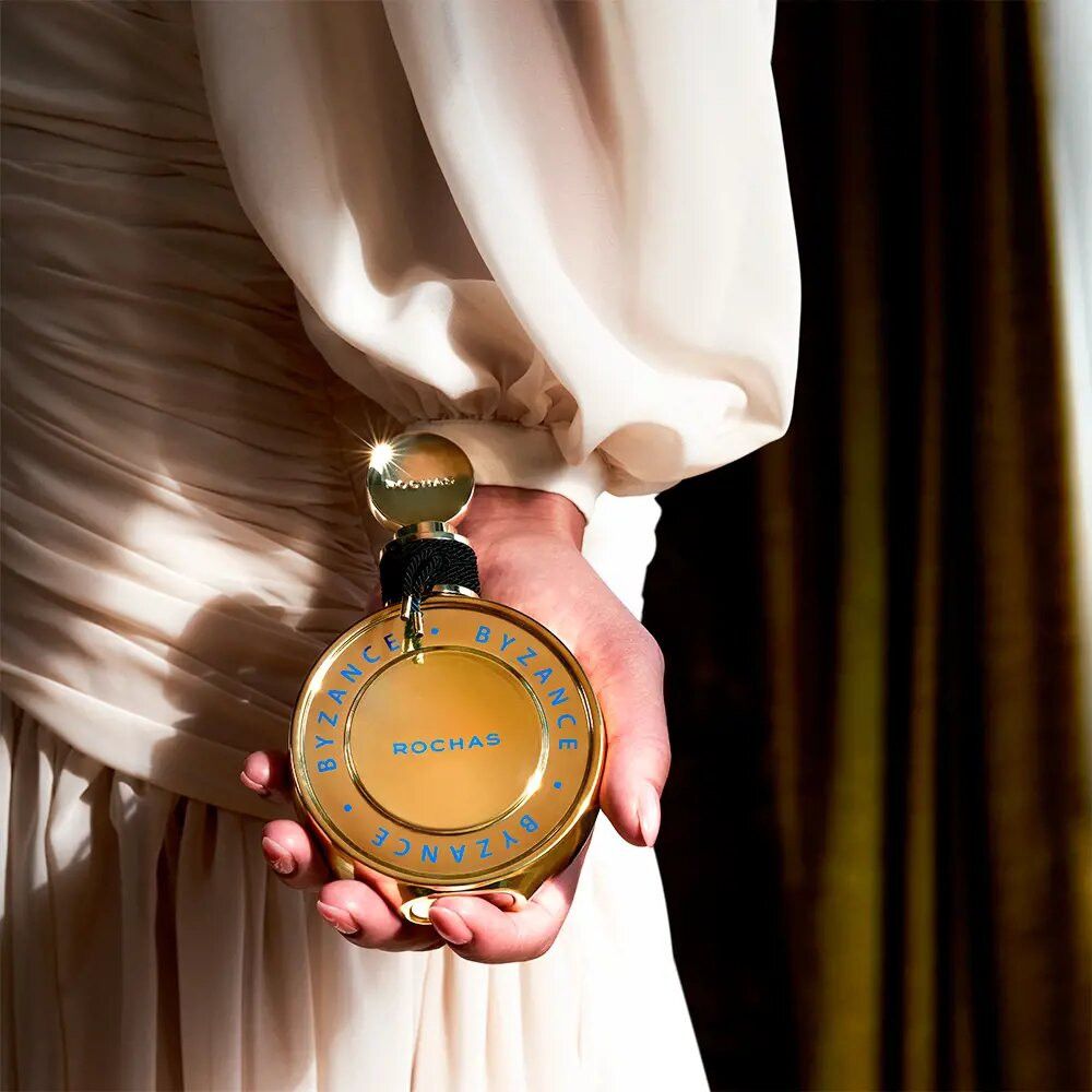 Flacon doré tenu par une personne. Inscription Byzance, Rochas. Cordon noir autour du col.