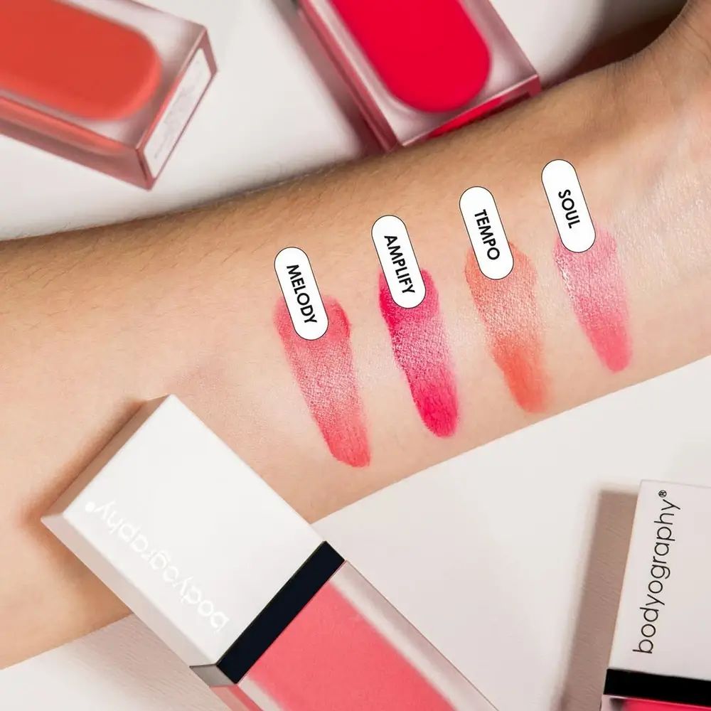 Arm met kleurstalen van BODYOGRAPHY vloeibare blush. Roze, oranje en rode tinten. Productverpakking.