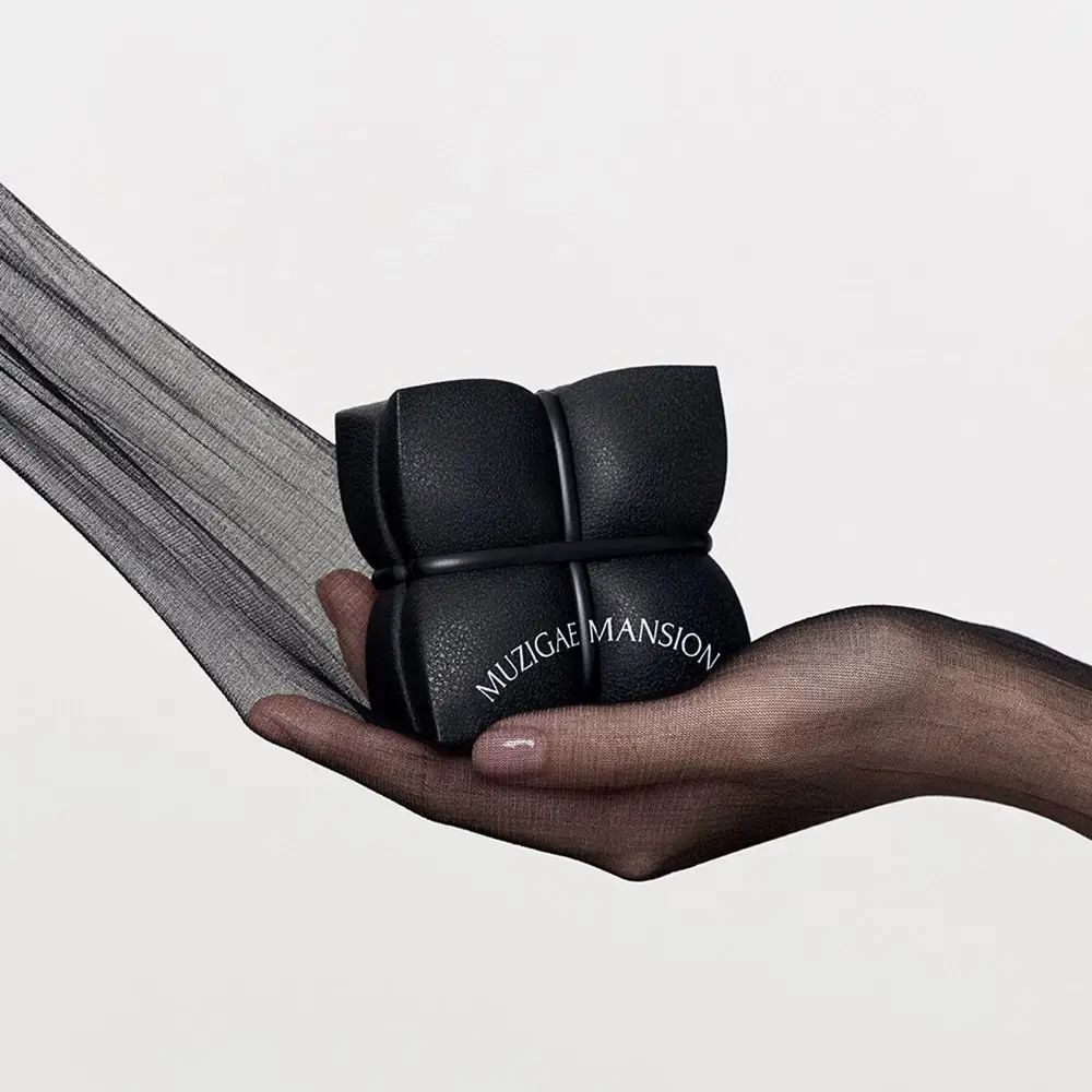 Hand houdt zwarte, vierkante etui met MUZIGAE MANSION-opschrift vast. Op de achtergrond zwarte stof.