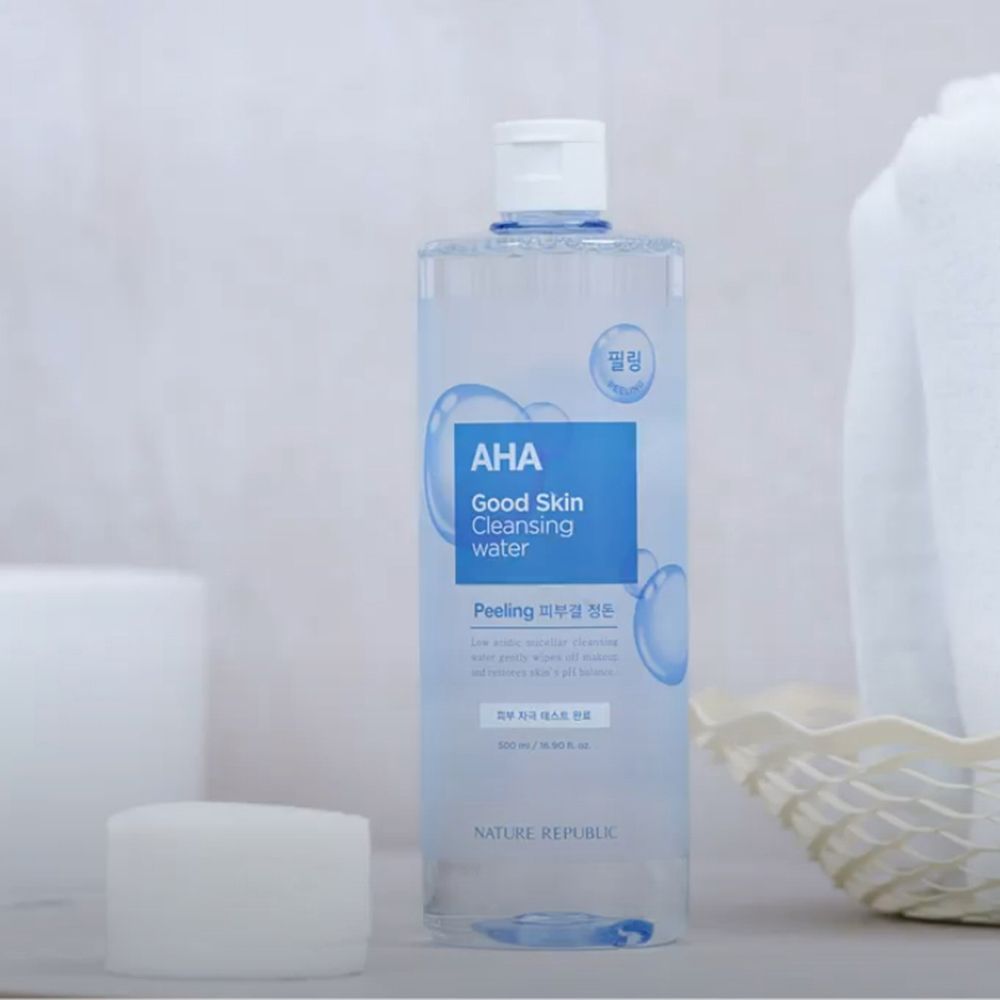 Flacon transparent avec étiquette bleue. Inscription: AHA Good Skin Cleansing Water. Marque: NATURE REPUBLIC. À côté d'une éponge blanche et d'une serviette.