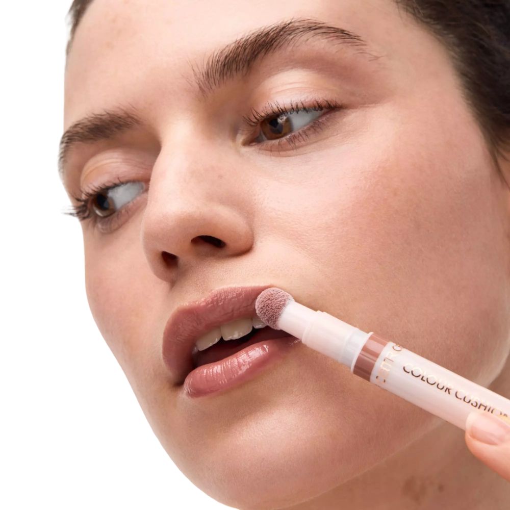 Persoon brengt lipolie aan. Roze applicator op lippen. Tekst: COLOUR CUSHION. Merk: CATRICE.