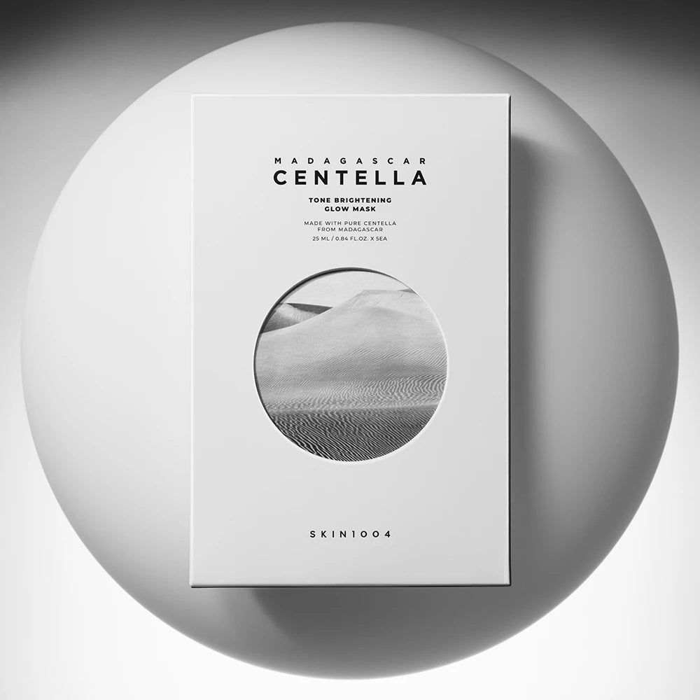 Witte productverpakking met tekst en ronde afbeelding. Opschrift: MADAGASCAR CENTELLA Tone Brightening Glow Mask. Merk: SKIN1004. Op witte achtergrond.