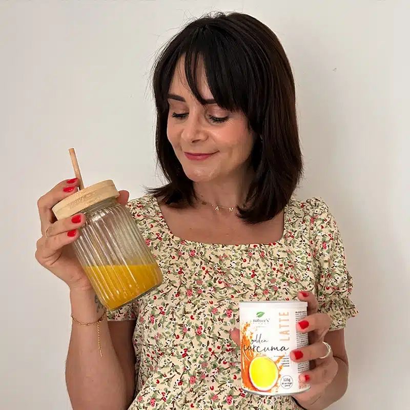 Vrouw houdt een glas met gouden drank en een blikje Nature's Finest Golden Curcuma Latte vast.