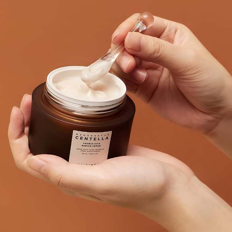 Mains tenant un pot de crème brun avec couvercle blanc et une spatule. Crème visible. Étiquette avec texte: MADAGASCAR CENTELLA, SKIN1004.