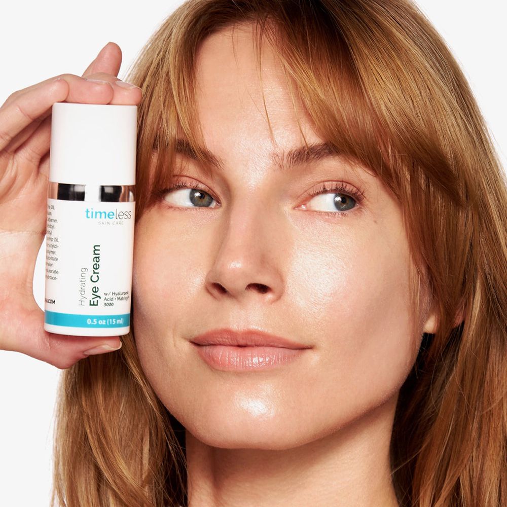 Een vrouw houdt een witte fles met zilveren ring en blauwe streep vast. Tekst: Hydrating Eye Cream. Merk: timeless skin care.