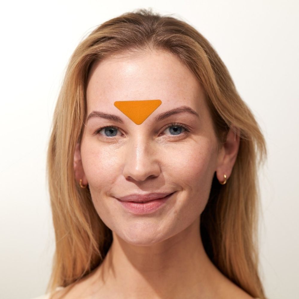 Vrouw met een oranje, driehoekige patch op het voorhoofd. Lichte huid en blond haar.