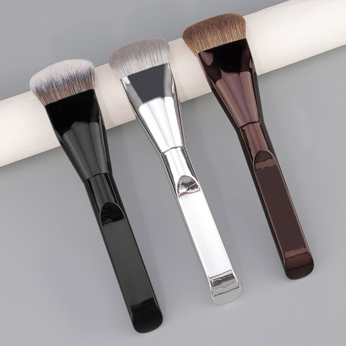 Trois pinceaux de maquillage plats avec manches noirs, argentés et marron et poils denses.