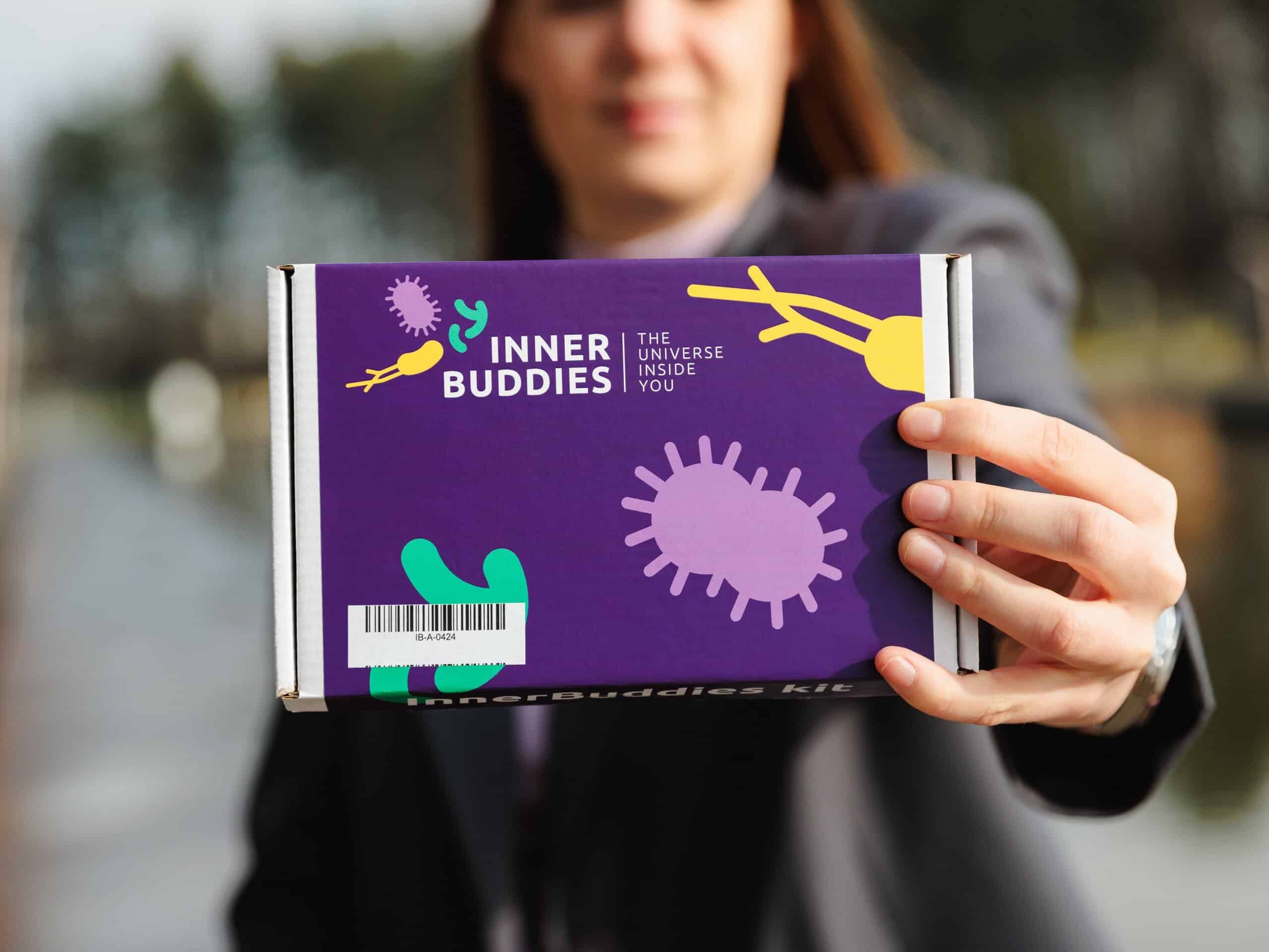 Une personne présente une boîte Inner Buddies sur fond violet avec des illustrations de microbes colorées.