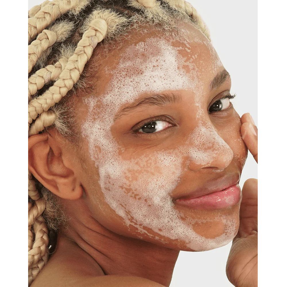 Visage d'une femme aux tresses blondes, recouvert de mousse. Elle se touche le visage.