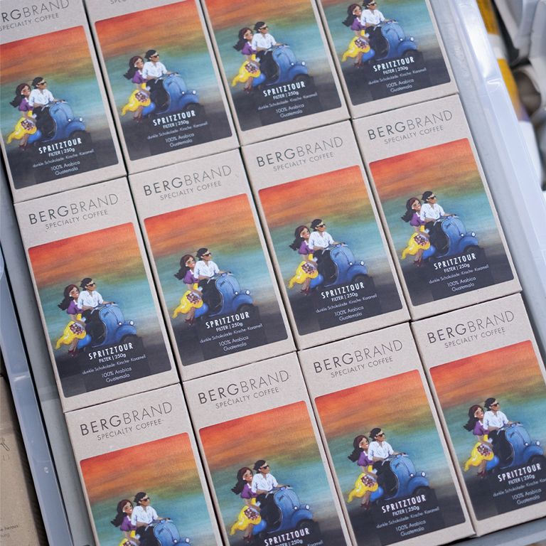 Plusieurs paquets de café BERGBRAND Spritztour. Boîtes avec illustration de personnes sur un scooter.