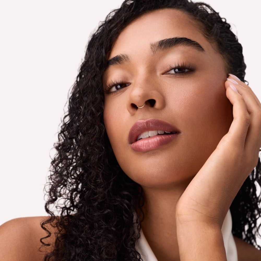 Portret van een vrouw met roze gloss op haar lippen. Ze raakt haar gezicht aan.