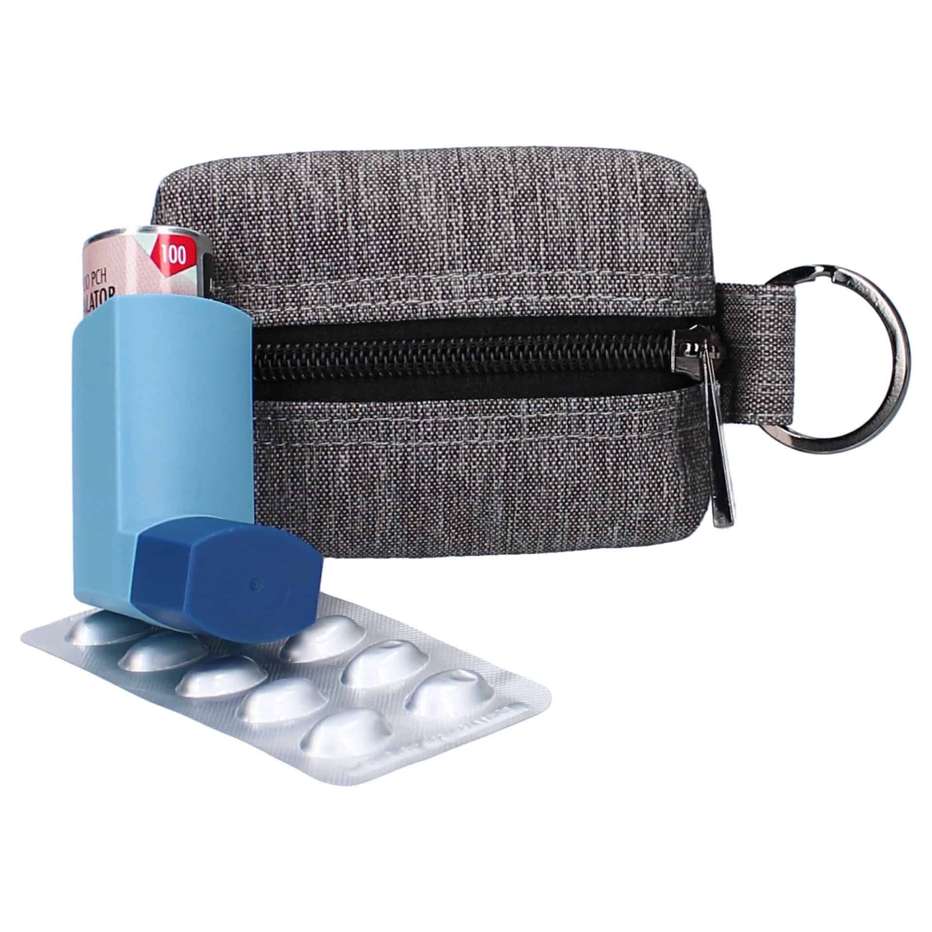Grijze stoffen etui met rits en sleutelring, ernaast een blauwe inhalator en een blisterverpakking met pillen.