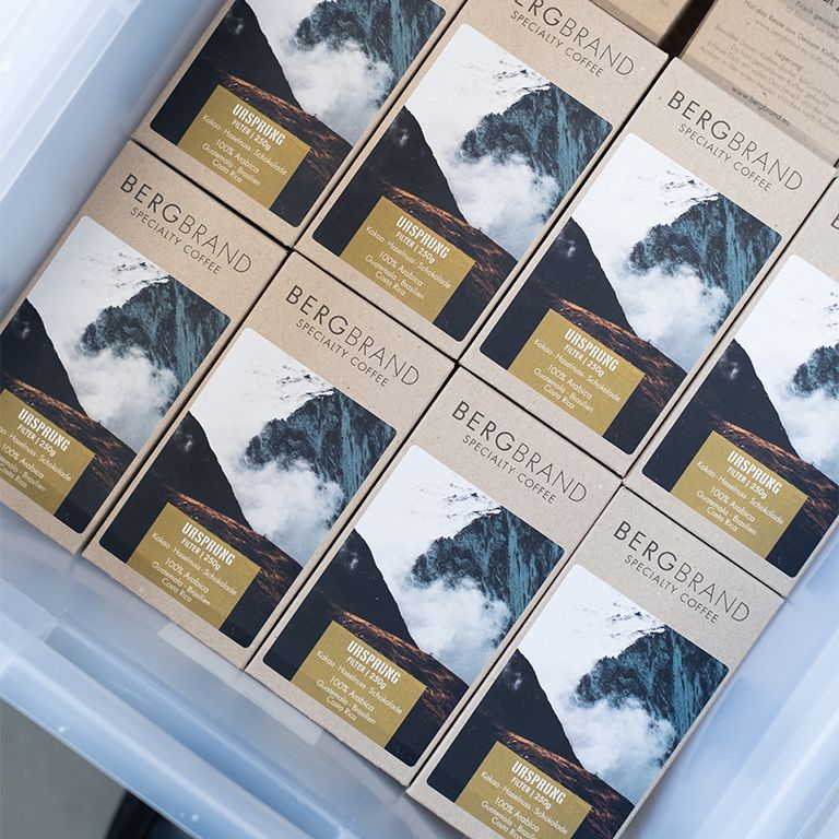 Plusieurs paquets de café avec motif de montagne. Texte : BERGBRAND Specialty Coffee, Ursprung Filter. Emballages dans une boîte transparente.