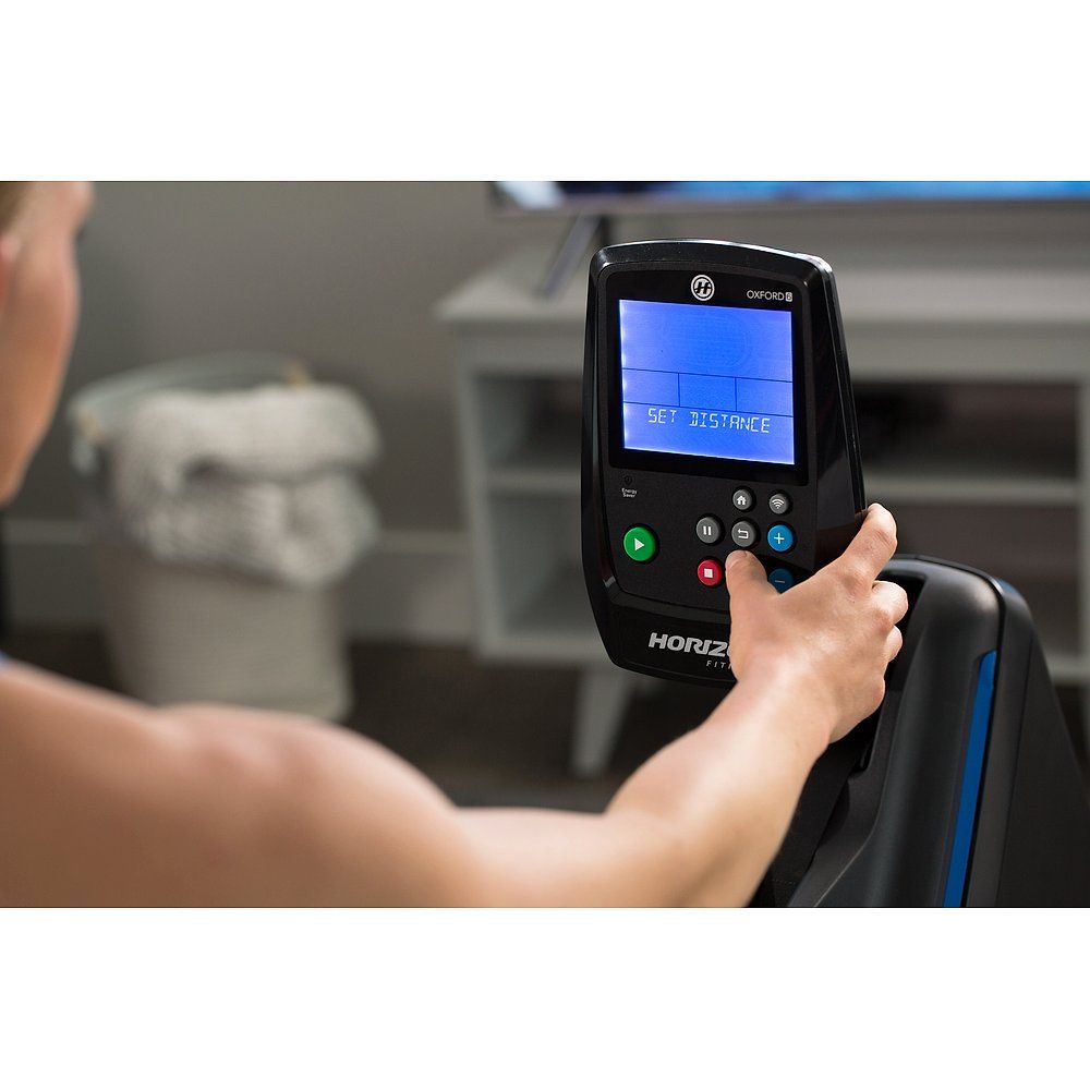 Persoon houdt bedieningspaneel vast. Display toont 'SET DISTANCE'. Horizon Fitness logo. Zwart paneel met knoppen.