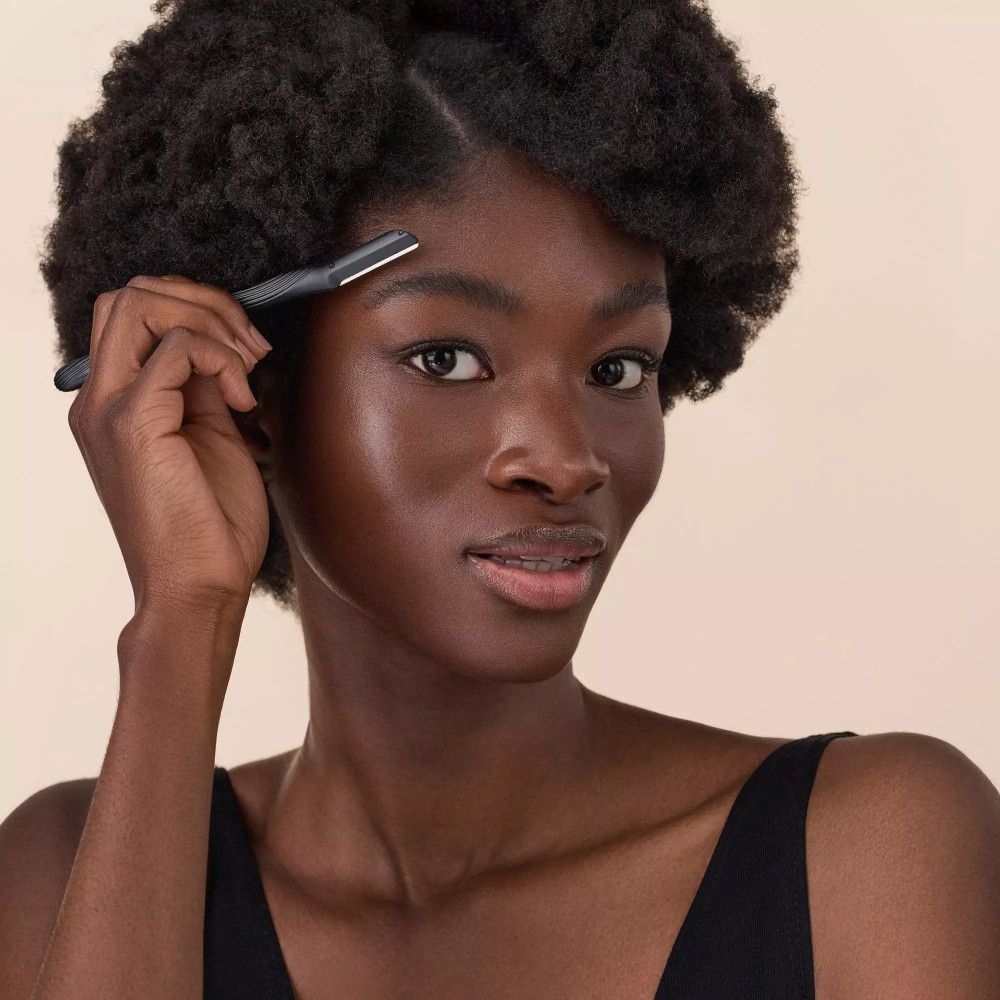 Une personne tient un rasoir à sourcils. Manche noir, lame argentée. Personne à la peau foncée et haut noir.