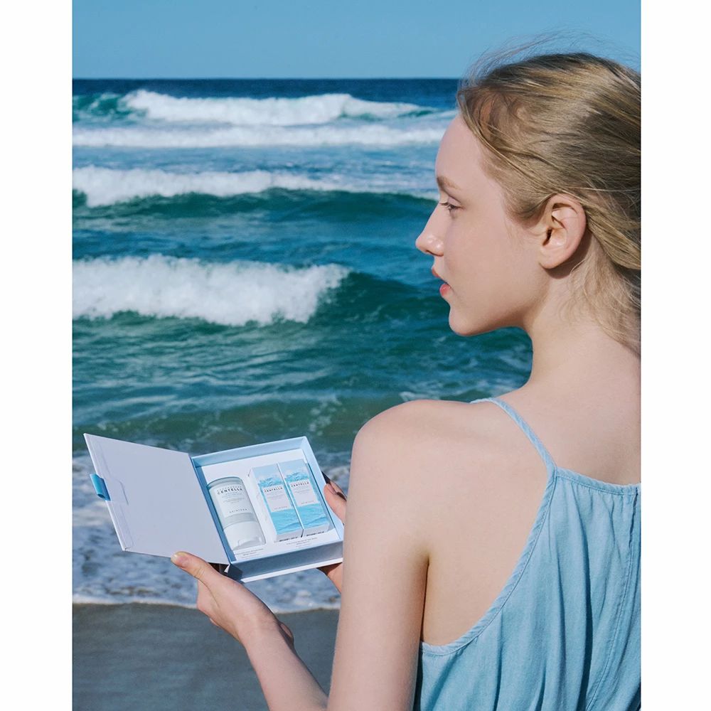 Vrouw houdt open productverpakking op strand vast. Bevat zonnebrandproducten. Achtergrond: zee en lucht.