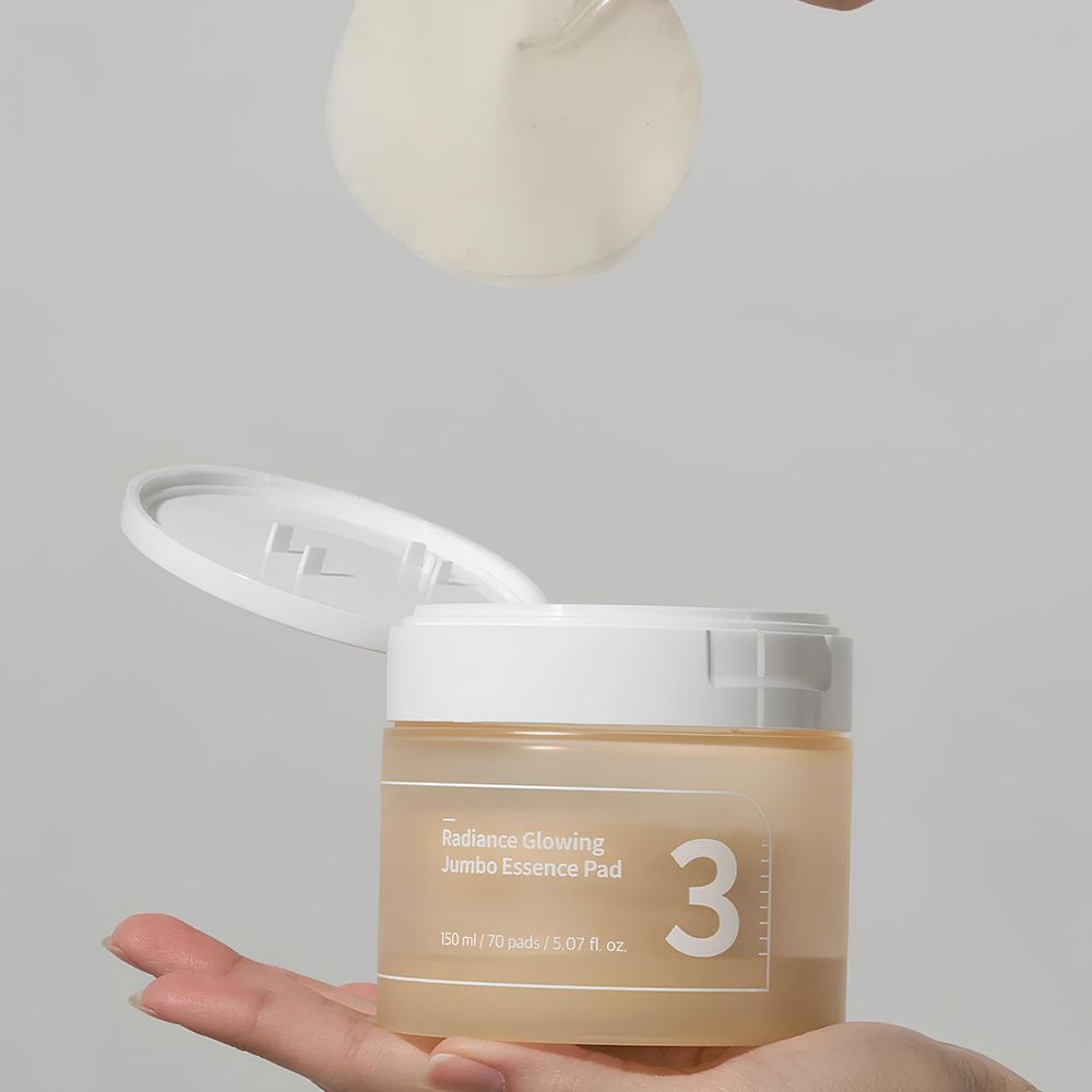 Pot beige avec couvercle blanc, ouvert. Inscription : 3, Radiance Glowing Jumbo Essence Pad. Un tampon est prélevé.