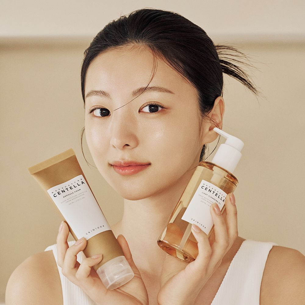 Une femme tient deux produits dans ses mains. À gauche, un tube de mousse ampoule, à droite, une bouteille d'huile nettoyante avec pompe. SKIN1004.