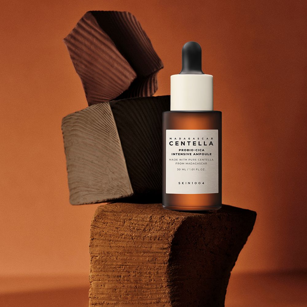 Bruine glazen fles met wit etiket. Zwarte pipet. Op houten blok. Tekst: CENTELLA, PROBIO-CICA, INTENSIVE AMPOULE, SKIN1004.