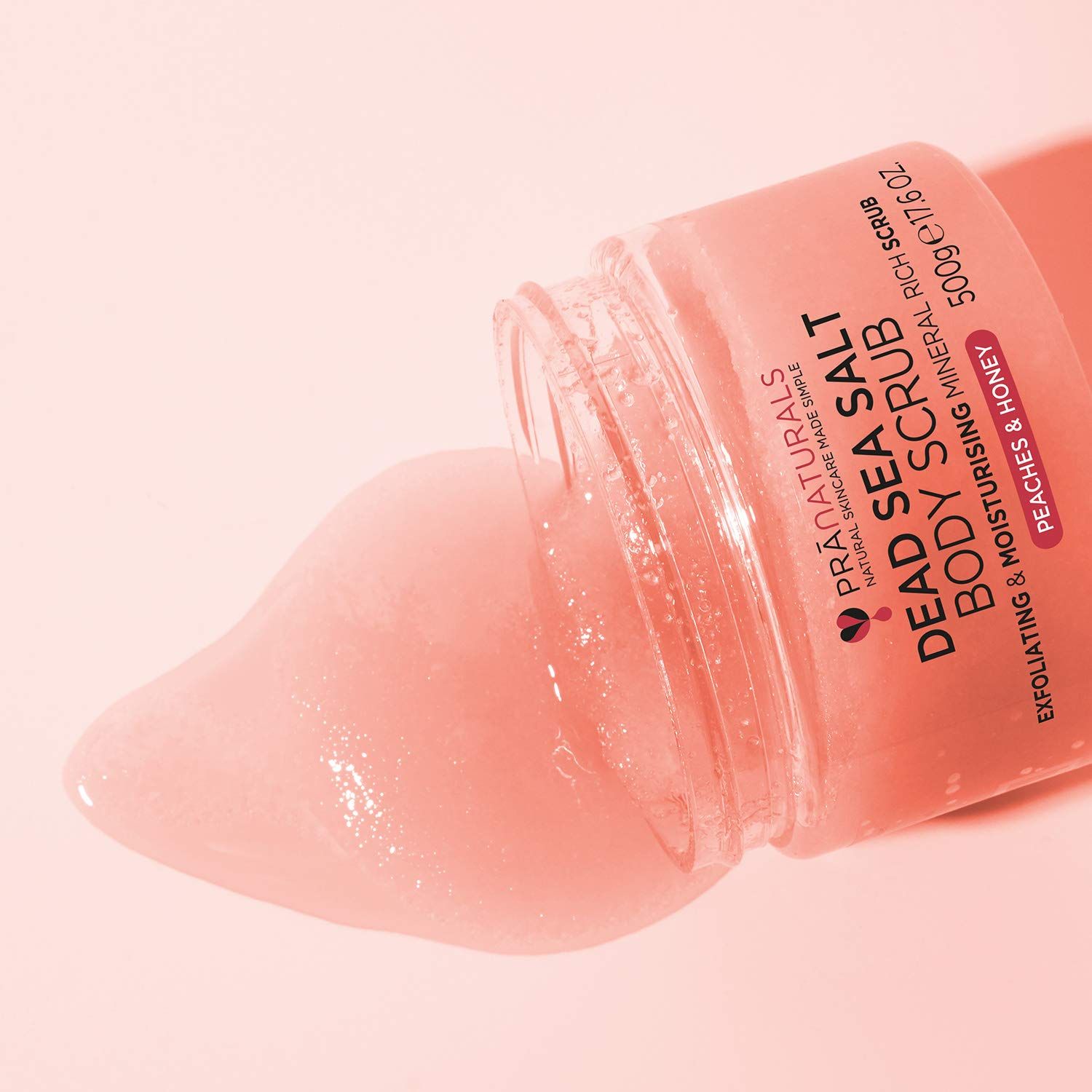 Pot bodyscrub schuin. Scrub vloeit eruit. Opschrift: Dead Sea Salt Body Scrub, Peaches & Honey. 500g/17.6 oz.