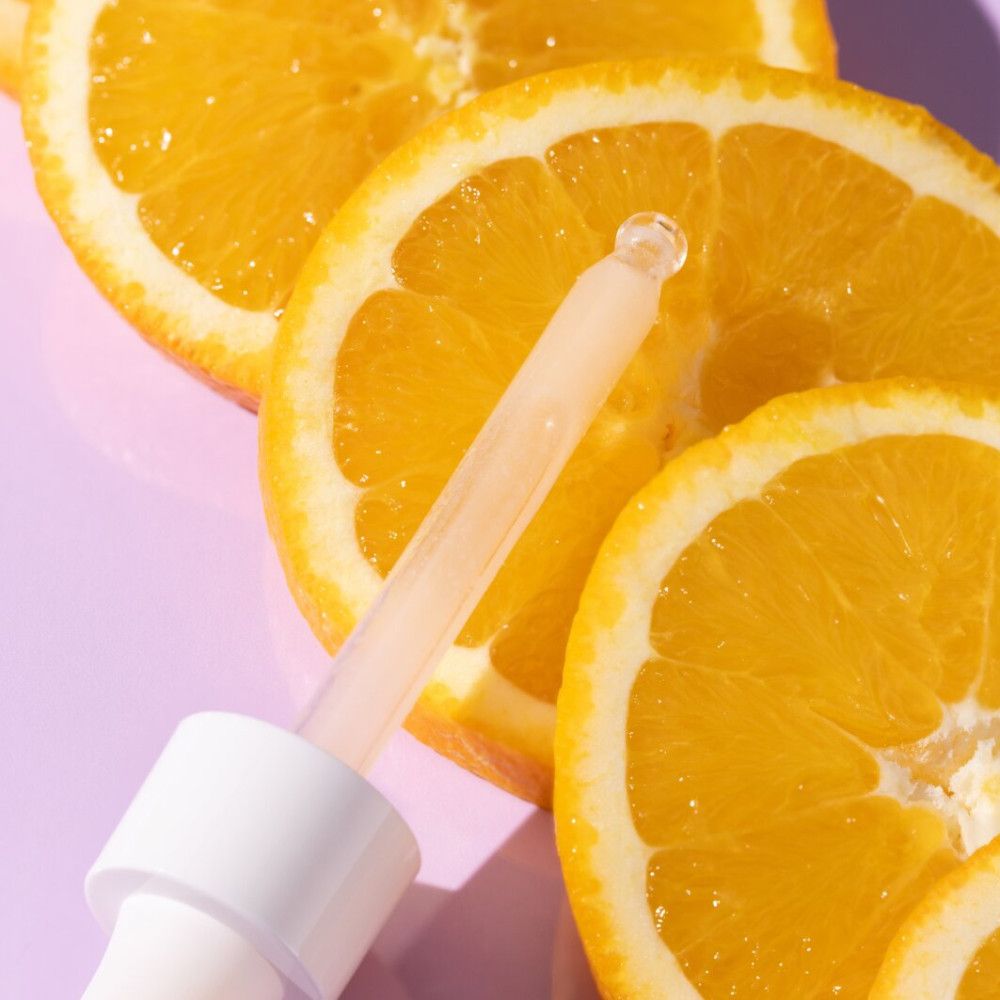 Compte-gouttes avec sérum devant des tranches d'orange. Le compte-gouttes est blanc, le sérum clair.