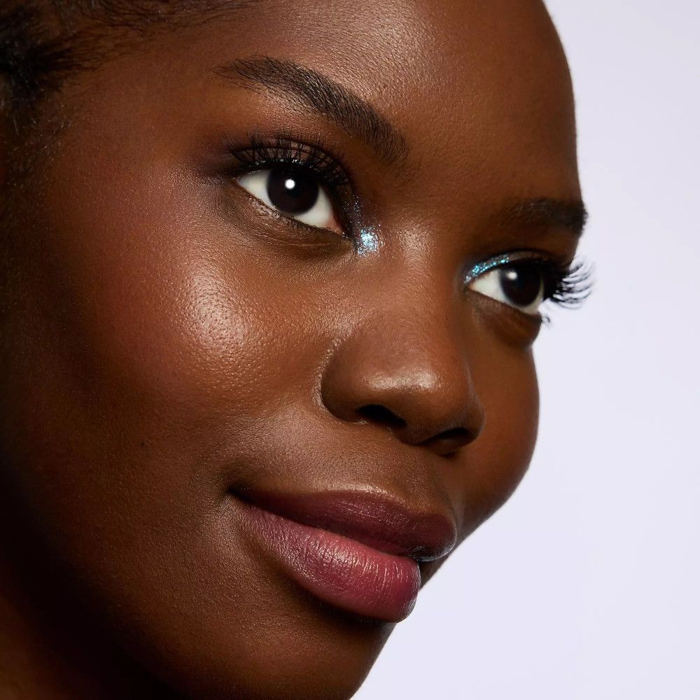 Gros plan d'un visage. Faux cils 3/4, peau foncée, peau brillante. Paillettes bleues sur l'œil.